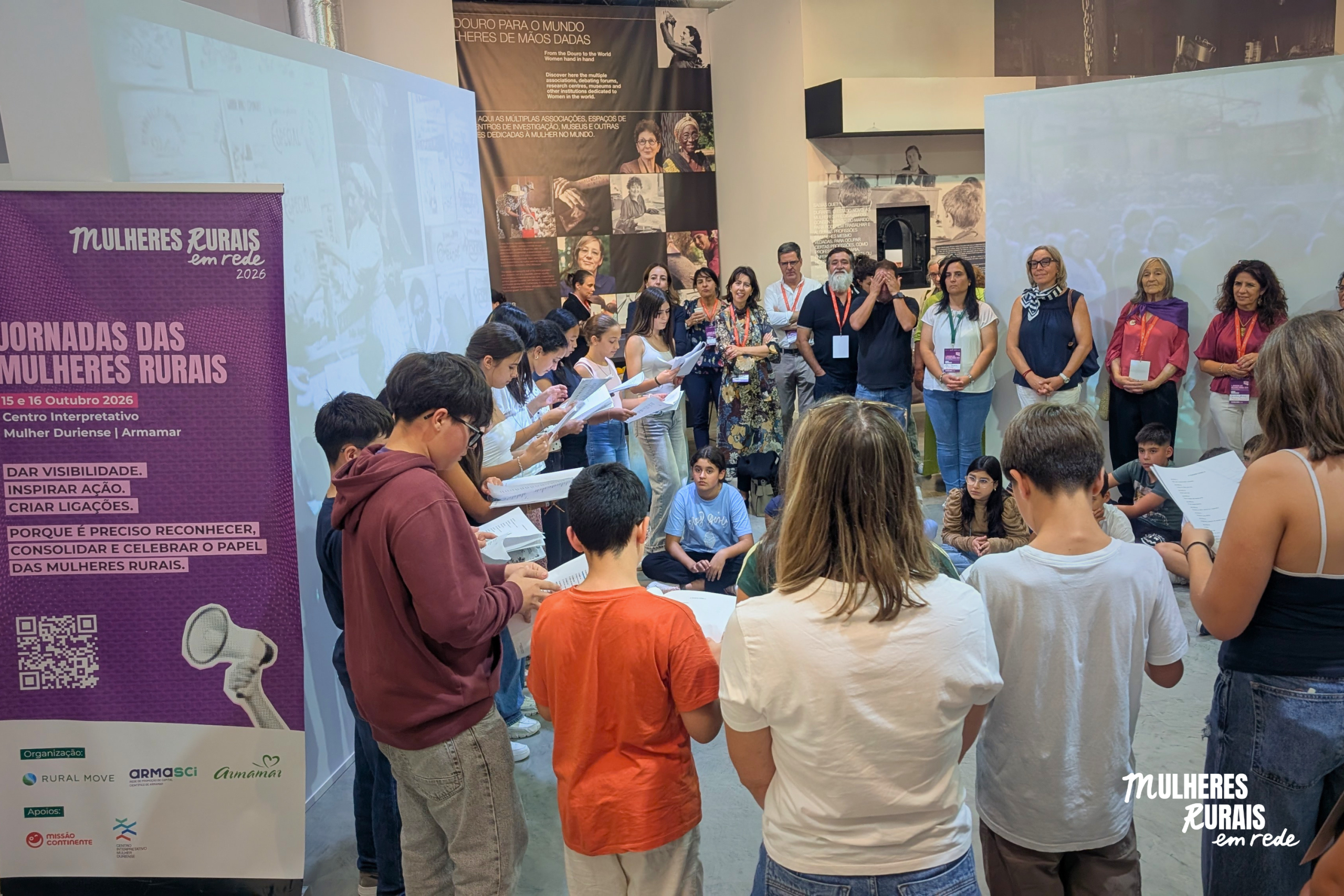 Jovens e participantes em roda, com folhas na mão, no Centro Interpretativo da Mulher Duriense, junto ao cartaz das Jornadas das Mulheres Rurais, em Armamar