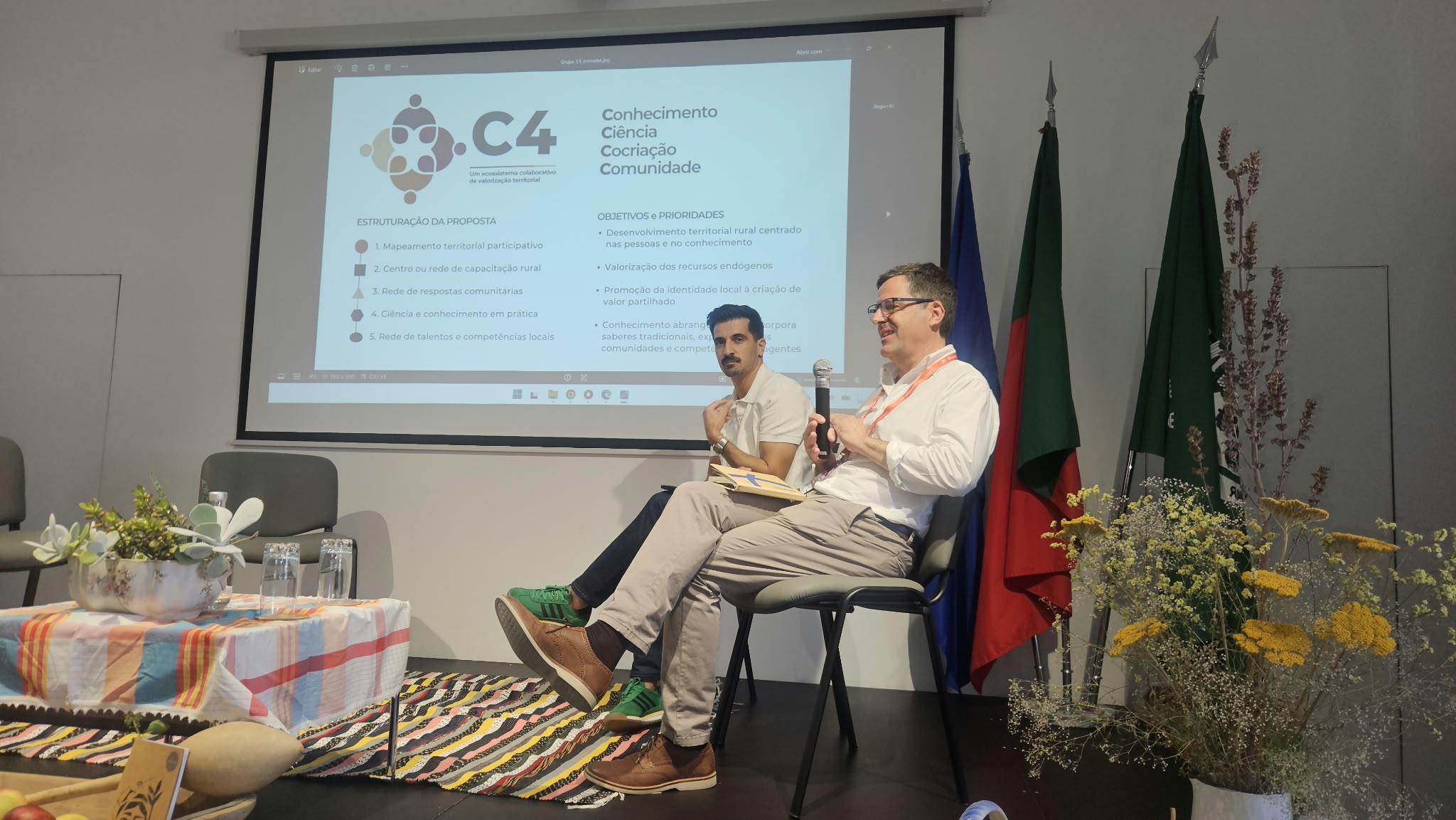 Dois intervenientes sentados em palco durante o momento de comentários aos projetos, com uma proposta de desenvolvimento rural projetada no ecrã