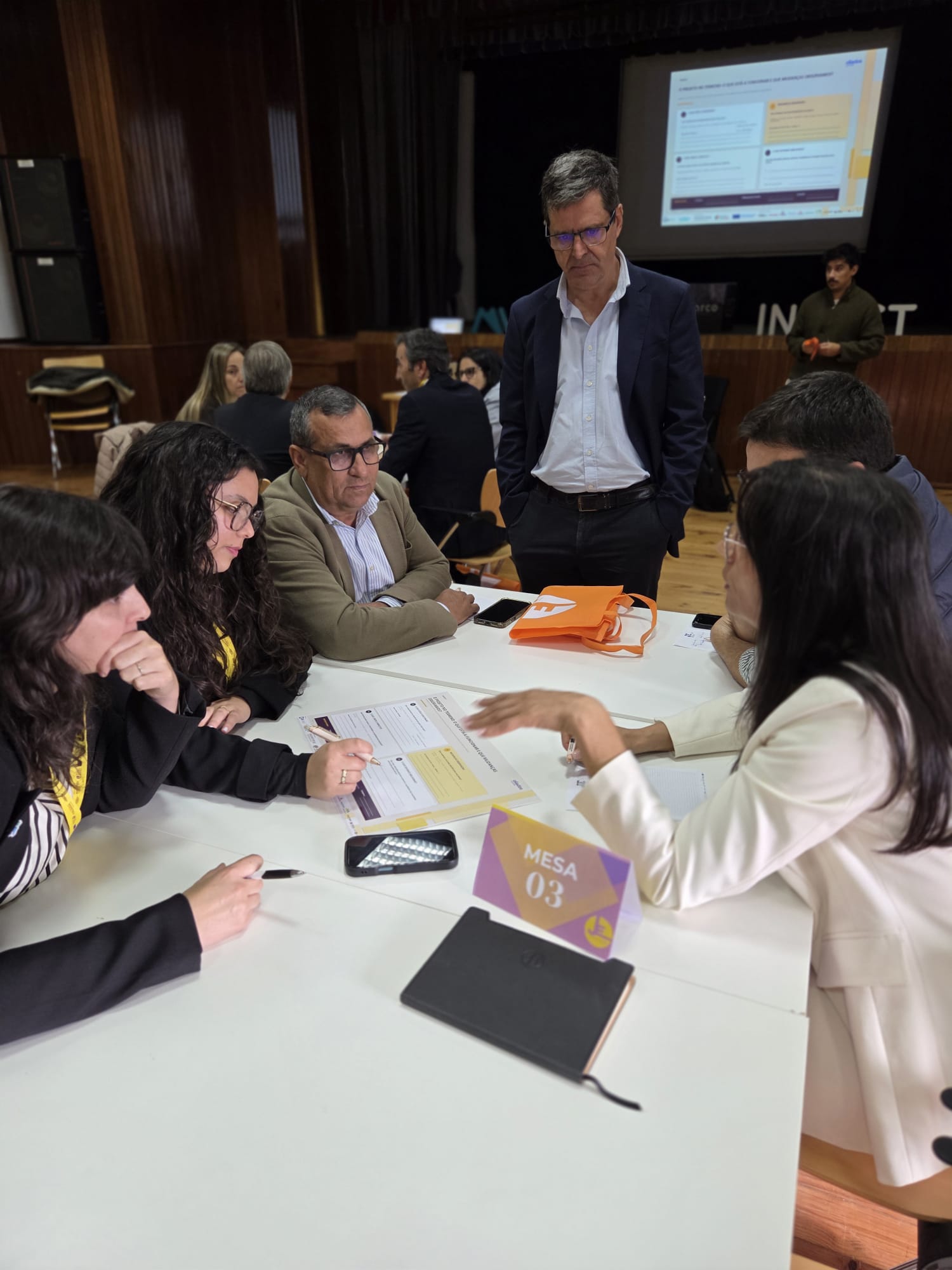 Participantes em trabalho de grupo à volta de uma mesa, com folhas de trabalho, durante a sessão de avaliação de impacto em Marco de Canaveses